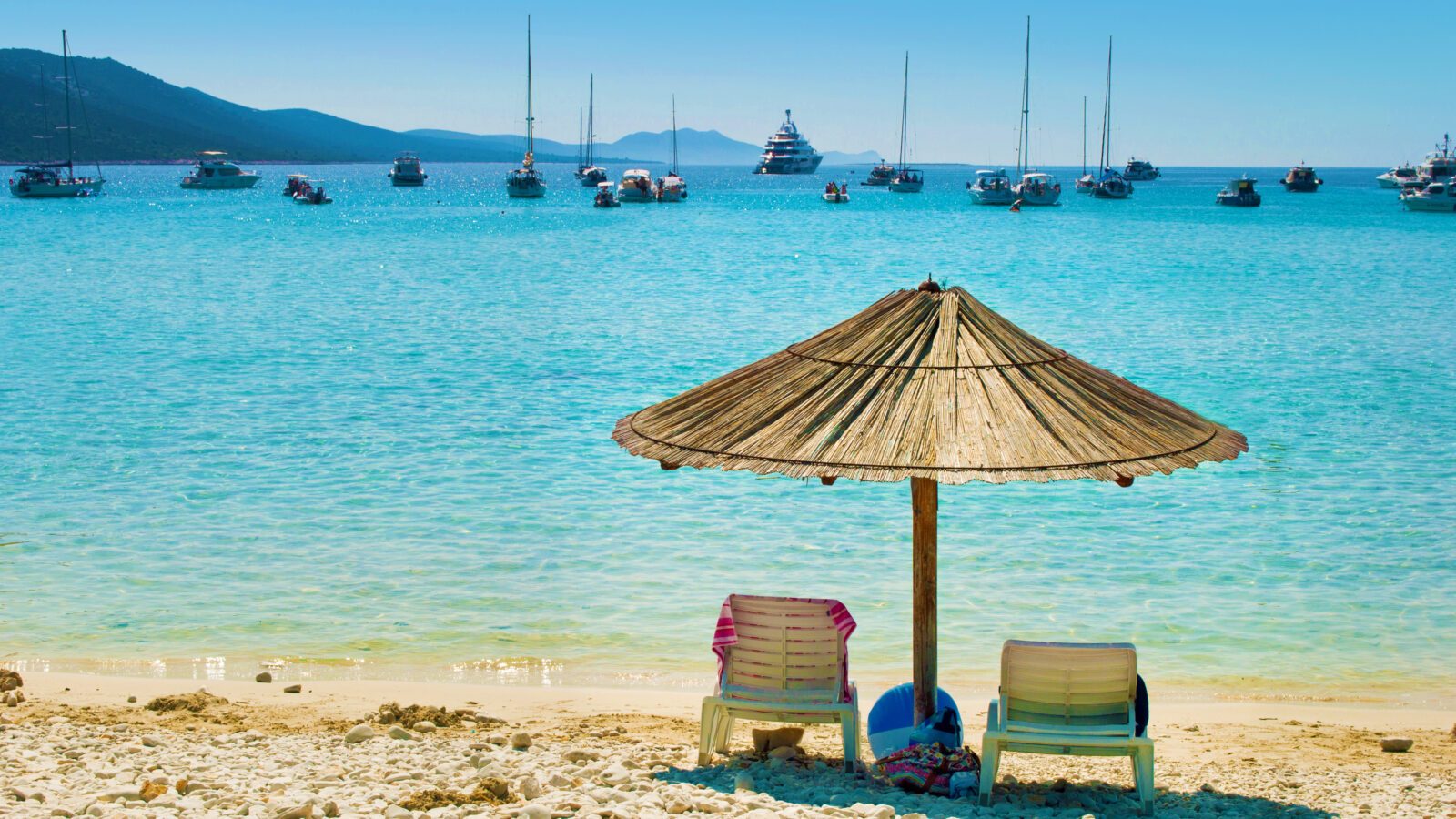 One yellow straw umbrella and two chairs on the pebble sandy beach. Many yachts in the azure blue sea lagoon in a bay near the coast of an island. Blue cloudless sky. Dugi otok, Croatia